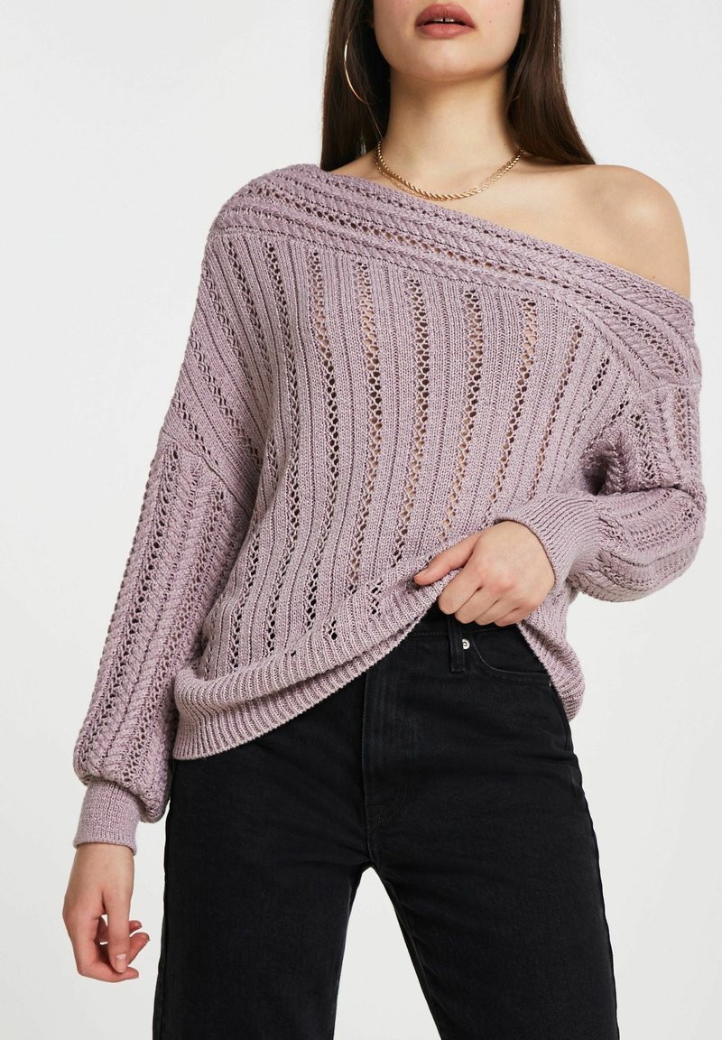 Pull tricoté violet clair avec texture côtelée et motif à mailles ouvertes. Encolure large décolletée et manches ballon. Porté sur un jean noir.