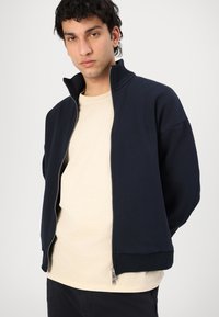 Marineblå zip-jakke med høy krage, ribbede mansjetter og avslappet passform, lagd over en lys beige T-skjorte. Glatt stofftekstur.