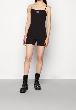 Černý úzký overal s tenkými ramínky a čtvercovým výstřihem, s malou nášivkou loga na hrudi. Modelka má na sobě černé šněrovací boty z krajky.