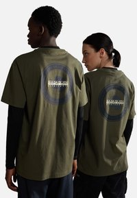 T-shirt in cotone verde oliva con un grande logo rotondo sulla schiena. Vestibilità casual, maniche corte, mostrati su due persone di spalle.