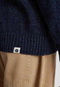 Dunkelblauer strukturierter Strickpullover mit geripptem Saum, versehen mit einem kleinen Stoffetikett mit Logo. Beigefarbene Hose mit dezenten diagonalen Streifen.