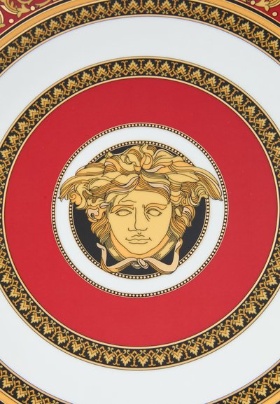 Kerámia tányér, melyen egy központi arany Medúza motívum látható piros háttér előtt, körülötte bonyolult fekete és arany virágminták.