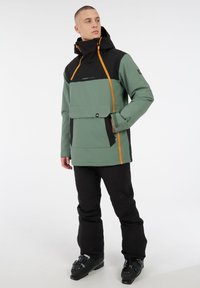 Giacca impermeabile verde e nera con una cerniera arancione, cappuccio, tasca frontale e maniche lunghe. Abbinata a pantaloni neri e stivali da sci.