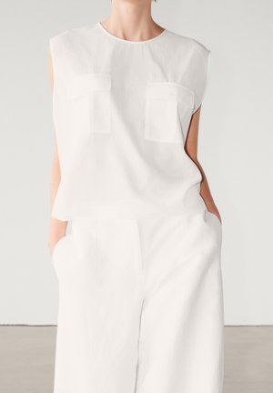 Femme portant un haut blanc sans manches avec deux poches poitrine et un pantalon blanc ample, les mains dans les poches, sur un fond uni.