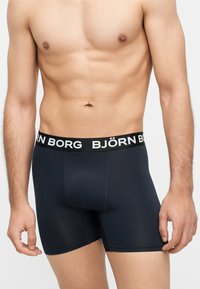 Αρσενικός κορμός που φοράει μαύρα μπόξερ Björn Borg με λουράκι με λογότυπο, στέκεται μπροστά σε μια απλή ανοιχτή επιφάνεια.