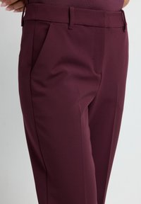 Pantalons taillés bordeaux avec une texture lisse, dotés de poches latérales et d'une taille ajustée, conçus pour une silhouette élancée.
