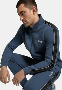 Tuta blu con zip caratterizzato da un alto colletto, strisce laterali nere a contrasto con loghi e tessuto leggermente testurizzato.