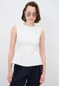 Weißes Peplum-Top mit ärmellosem Design, glattem Stoff und dezenten Nähten, kombiniert mit navyfarbener Hose und reflektierenden Sonnenbrillen.