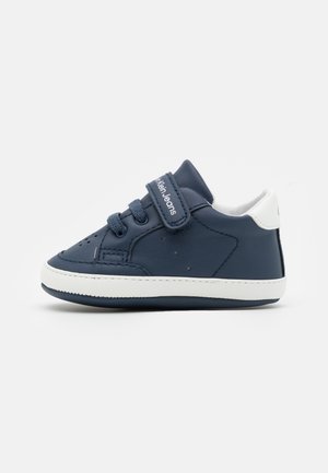 Baskets bleu marine pour tout-petit avec languette de talon blanche, semelle blanche, lacets et une bride Velcro portant la mention Calvin Klein Jeans sur un fond uni.