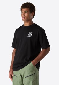 Mladý muž v černém tričku s malým bílým kresleným logem na hrudi a světle zelených cargo kalhotách se zipovými kapsami.