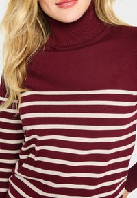 Femme blonde portant un pull à col roulé bordeaux avec des rayures blanches horizontales sur la poitrine et les manches.