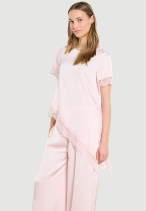 Young woman wearing pale pink asymmetrical lace-trimmed top and matching wide-leg pants, standing against a plain light background.