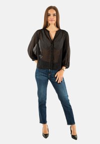 Blusa negra semitransparente con botones y mangas largas abullonadas, llevada sobre unos vaqueros azules y tacones negros, que muestra un ajuste holgado y una textura ligera.