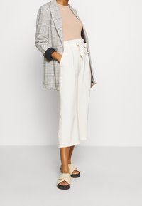 Veste grise à carreaux portée sur un haut côtelé beige ; pantalon blanc taille haute raccourci avec un détail nœud ; sandales beige à enfiler.