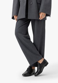 Scarpe di pelle nera con una finitura liscia, dotate di dettagli con lacci; indossate con pantaloni grigi larghi e un blazer grigio oversize.