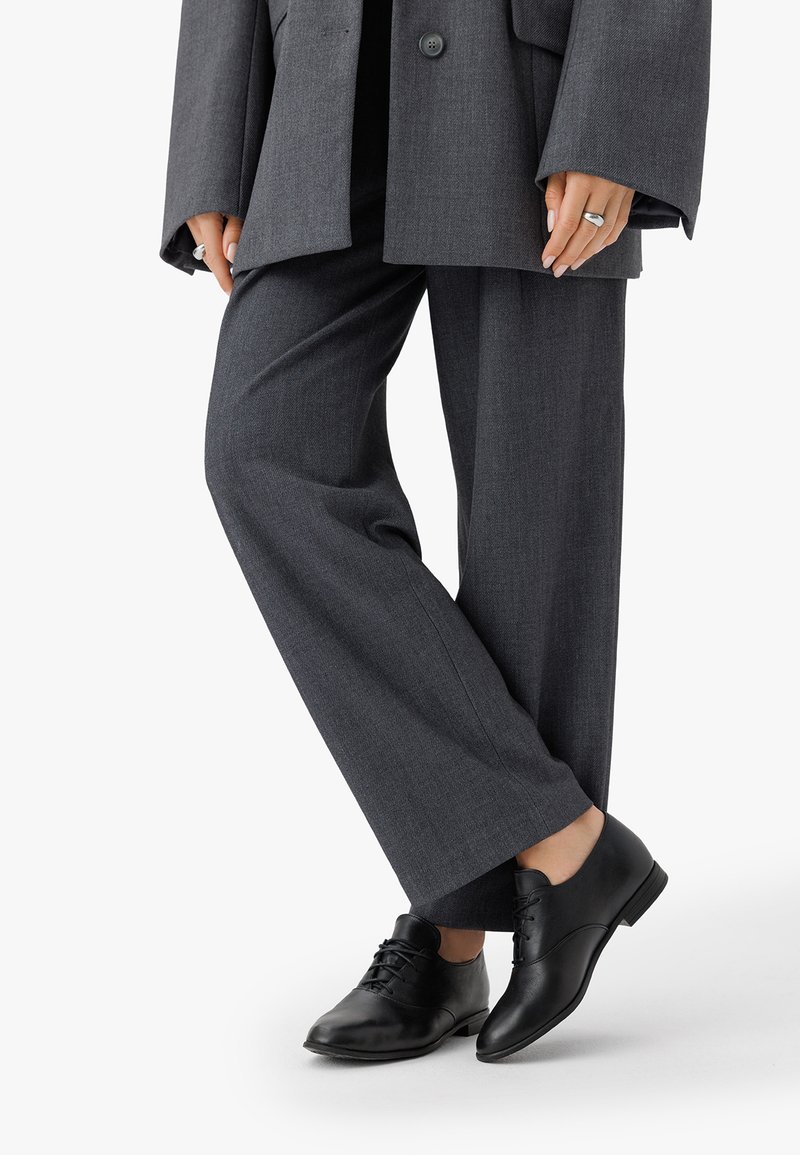 Scarpe di pelle nera con una finitura liscia, dotate di dettagli con lacci; indossate con pantaloni grigi larghi e un blazer grigio oversize.