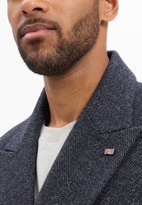 Marineblauwe herringbone overjas met grote revers, gestructureerde stof en een kleine Britse vlagspeld op de kraag, gedragen over een lichte trui.