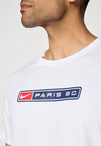 Hvid bomulds T-shirt med et blåt rektangulært design, der har "PARIS SG" og et rødt Nike-logo, rund halsudskæring, korte ærmer.