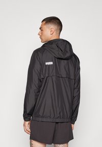 Chaqueta ligera negra con capucha y cintura elástica, con un logo en la parte posterior. Tejido suave con un diseño minimalista.