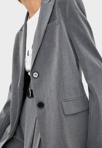 Person som bär en grå skräddarsydd blazer med två knappar över en vit skjorta med tryck och matchande grå byxor med ett svart bälte.