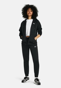 Zwarte hoodie met een ritssluiting, zijzakken en een wit logo. Gecombineerd met zwarte joggingbroeken en zwart/witte sportschoenen.