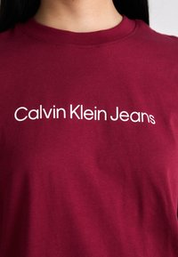 T-shirt en coton bordeaux avec texte blanc à l'avant : "Calvin Klein Jeans." Encolure classique à crew avec une texture douce et lisse.