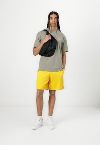 T-shirt gris avec logo, shorts jaunes, baskets blanches et sac en bandoulière noir. Le modèle se tient devant un fond blanc uni.