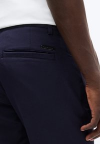 Κοντινό πλάνο του χεριού ενός ατόμου που ακουμπά κοντά στην πίσω τσέπη μπλε ναυτικού χρώματος παντελονιού με μια μικρή μαύρη μεταλλική ετικέτα λογότυπου.