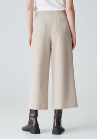 Pantaloni beige a gamba larga con una texture liscia, due tasche posteriori e vita alta, abbinati a stivali con tacco marroni.
