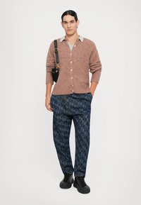 Cardigan tricotat maro cu textură în bobble, guler gri, pantaloni întunecați cu imprimeu și adidași negri. Ținută casual cu o geantă de umăr.