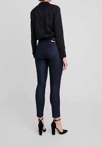 Pantalones ajustados de mezclilla de color azul oscuro, con una textura suave y bolsillos traseros. Combinados con una camisa negra con cuello y zapatos de tacón negros.