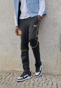 Persoon met zwarte Puma sneakers en zwarte trainingsbroek met gele piping en het Borussia Dortmund-logo, staande op een klinkerstraat.