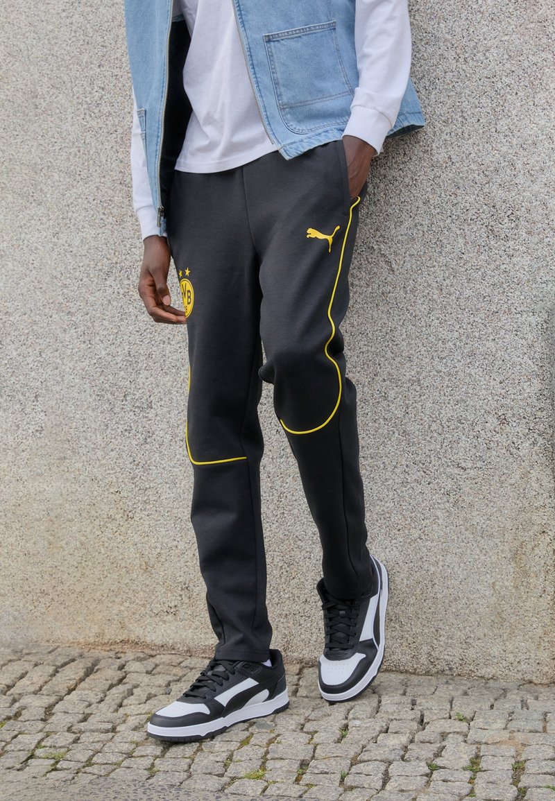 Persoon met zwarte Puma sneakers en zwarte trainingsbroek met gele piping en het Borussia Dortmund-logo, staande op een klinkerstraat.