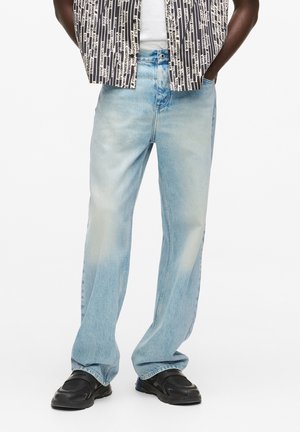 Homme portant un jean droit délavé bleu clair, des baskets noires, une chemise blanche et une chemise ouverte à motif noir et blanc.