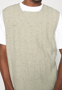 Suéter sin mangas de punto en gris claro con textura de canalé vertical y sutiles motas. Llevado sobre una camisa blanca de manga corta.