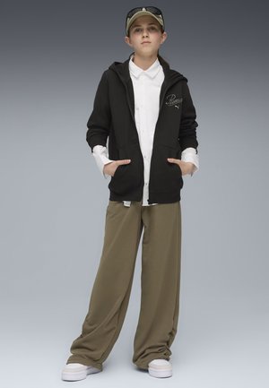 Schwarze Zip-Hoodie mit Fronttaschen, darunter ein weißes Hemd mit Kragen, olivgrüne, lässige Hose und weiße Sneakers, ergänzt durch eine hellgrüne Kappe.