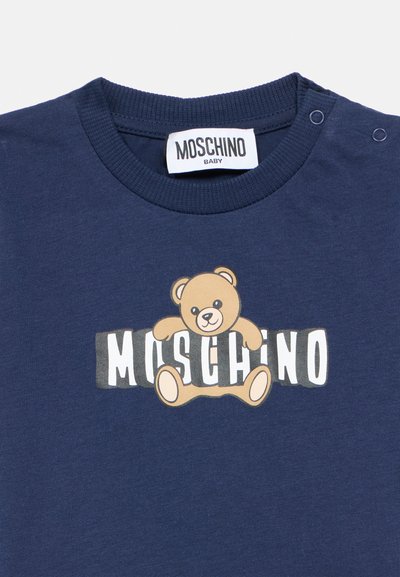 Μπλε ναυτικό βαμβακερό μπλουζάκι με σχέδιο ενός αρκουδάκου και το κείμενο «Moschino» σε λευκό και γκρι. Διαθέτει κλείσ ιμο με κουμπιά στον ώμο.