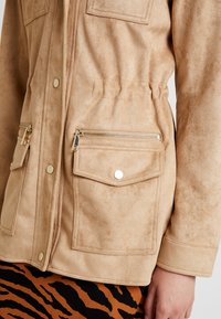 Giacca in suede color beige con una superficie leggermente testurizzata. Presenta due tasche con zip e chiusure a pressione. Progettata con una vita regolabile.