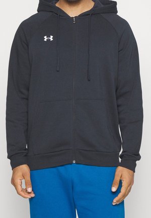 Mann trägt einen schwarzen Zip-Hoodie mit einem weißen Under Armour-Logo und hellblauen Hosen, steht vor einem schlichten Hintergrund.