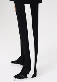 Pantalon noir avec une rayure verticale blanche, fabriqué en tissu doux. Associé à des baskets noires avec des détails de couture blancs.
