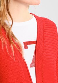 Czerwona, dziana kardiganka o teksturowym wzorze, z otwartym przodem, noszona na białym T-shircie z błyszczącą czerwoną cyfrą 7.