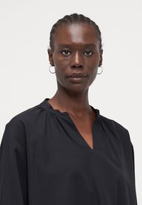 Black blouse with a relaxed fit, featuring a V-neckline and gathered detail at the collar. Smooth texture with a lightweight fabric.