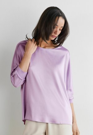 Frau mit schulterlangem dunklem Haar trägt ein locker sitzendes lavendelfarbenes Langarmshirt und beige Hosen, blickt nach unten, die Hand nahe am Hals.