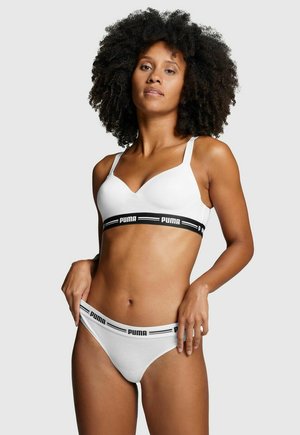 Frau mit natürlichem lockigem Haar, die einen weißen Puma-Sport-BH und passende Unterwäsche mit schwarzen elastischen Bändern mit Markenlogo trägt, vor einfarbigem Hintergrund posiert.