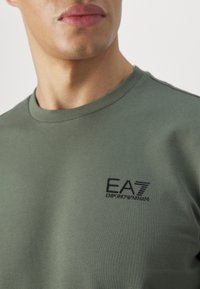 T-shirt verde oliva in cotone, con collo a girocollo e un piccolo logo EA7 ricamato nero sul petto. Texture morbida e design aderente.