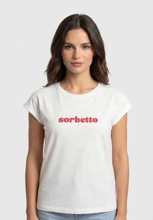 Mujer con cabello largo y oscuro, vistiendo una camiseta blanca con la palabra "sorbetto" en texto rojo, de pie frente a un fondo claro y sencillo.