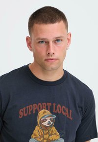 Camiseta de algodón azul marino con un gráfico de un perezoso en una sudadera amarilla y el texto "APOYA LO LOCAL" en letras negritas de color marrón rojizo.