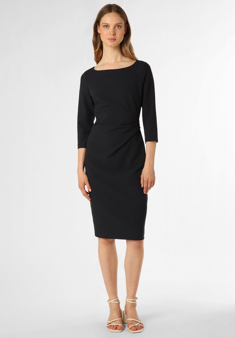 Schwarzes, eng anliegendes Kleid mit quadratischem Ausschnitt, Dreiviertelärmeln und einem verdrehten Taillendesign. Model trägt hautfarbene, geschnürte High Heels.