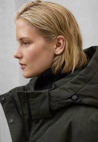 Tmavě zelená bunda s černým svetrem s vysokým límcem pod ní. Má přední zip, zapínání na druky a strukturovaný límec.