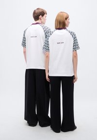 Λευκά T-shirts με σχεδιασμένα κοντά μανίκια σε τιρκουάζ και μαύρο, με μωβ γιακά και το κείμενο "EST. 1978" στην πλάτη. Συνδυασμένα με μαύρο παντελόνι με φαρδιά πόδια.